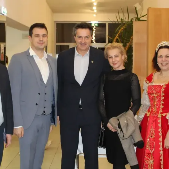 Fünf Menschen, drei Männer und zwei Frauen, posieren für ein Foto in einem Raum. Zwei Frauen tragen Kleider, eine mit einer Krone. Die Männer tragen Anzüge und Krawatten.