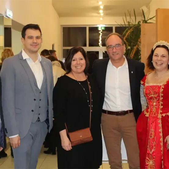 Fünf Menschen stehen in einer formellen Umgebung zusammen. Zwei Männer tragen Anzüge, einer mit Weste. Eine Frau in Rot und Gold steht zwischen zwei Männern, alle lächeln. Eine Pflanze und ein Schild sind im Hintergrund.