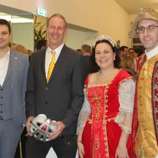 Drei Männer und eine Frau stehen lächelnd zusammen. Ein Mann hält einen Helm. Die Frau trägt ein rotes Kleid mit Goldbesatz. Die Männer tragen Anzüge und Krawatten, einer mit einer gelben Krawatte. Sie scheinen sich auf einer formellen Veranstaltung zu befinden.