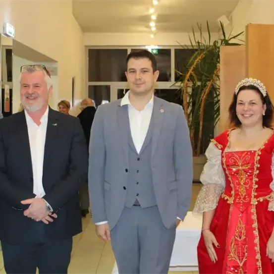 Fünf Menschen in formeller Kleidung stehen lächelnd zusammen in einem Raum. Der zentrale Mann trägt einen grauen Anzug, während die Frau in Rot mit Krone und Halskette geschmückt ist.