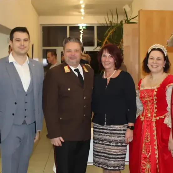 Eine Gruppe von Menschen steht lächelnd für ein Foto in einem Raum. Ein Mann in Uniform und eine Frau in einem roten Kleid stehen in der Mitte.