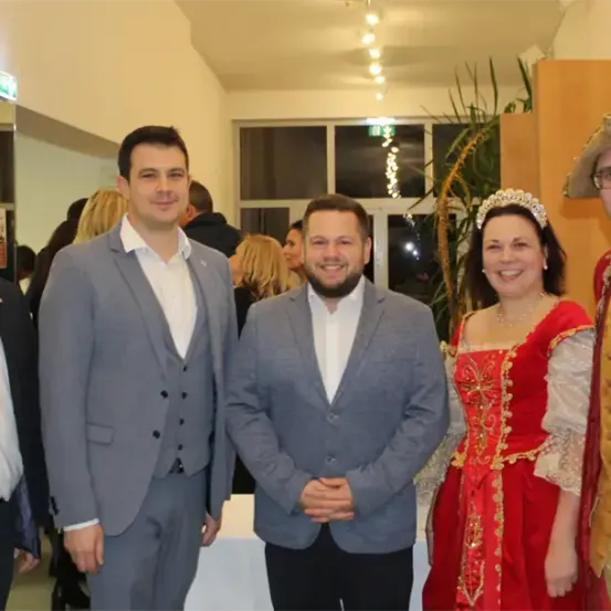 Fünf Personen, darunter zwei Männer in Anzügen und eine Frau in einem roten Kleid, posieren für ein Foto. Dahinter stehen zwei weitere Personen in der Nähe eines weißen Tisches.