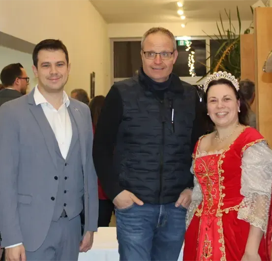 Fünf Menschen stehen in einem Raum und lächeln für ein Foto. Ein Mann in einem Anzug steht links, gefolgt von einem Mann in einer schwarzen Weste. Daneben steht eine Frau in einem roten und weißen Kostüm. Dahinter steht ein Mann in einem goldenen Hut.