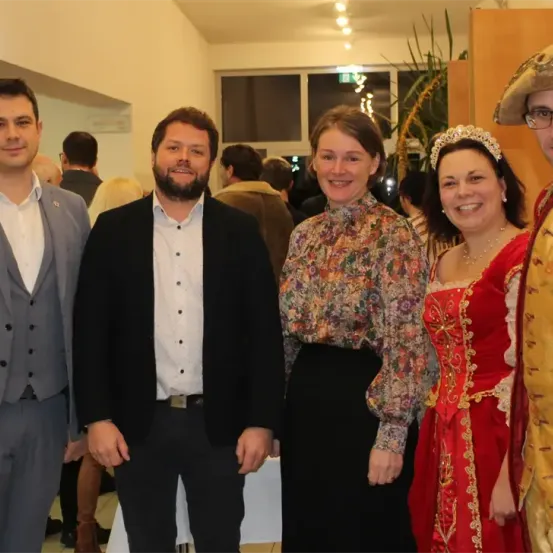 Eine Gruppe von Menschen lächelt und posiert für ein Foto. Zwei Frauen in der Mitte tragen historische Kostüme. Hinter ihnen stehen und sitzen Menschen an einem Tisch.