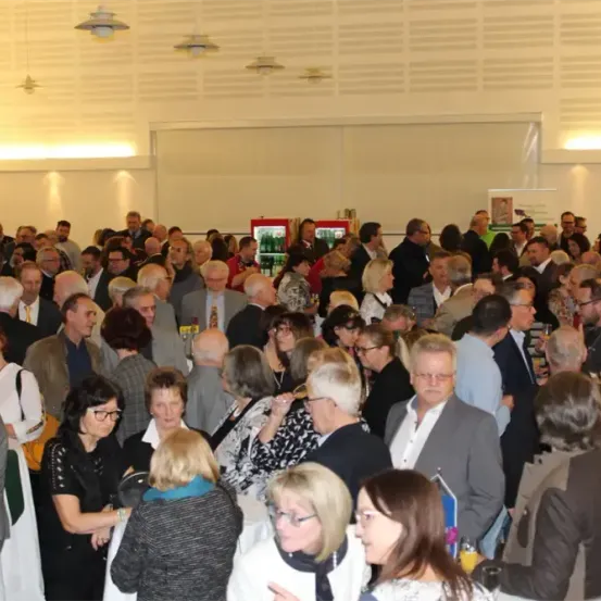 Eine große Menschenmenge in formeller Kleidung versammelt sich drinnen, einige stehen, andere unterhalten sich, wobei ein Kühlschrank und Lampen im Hintergrund zu sehen sind.
