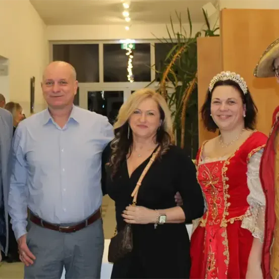 Vier Menschen posieren für ein Foto in einem Raum mit festlicher Atmosphäre. Zwei Frauen sind in aufwendigen Kostümen gekleidet, während die Männer formelle Kleidung tragen. Die Frau links trägt ein schwarzes Kleid und eine Halskette.