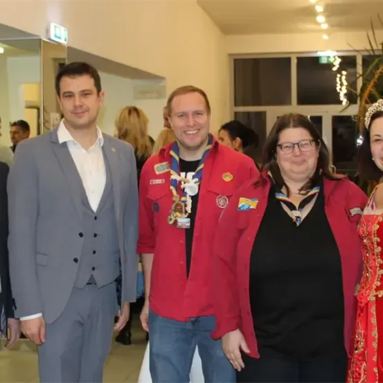 Eine Gruppe von Erwachsenen, darunter zwei Frauen und drei Männer, posiert für ein Foto in einem Raum. Die Männer tragen formelle Kleidung, während die Frauen eine Krone und ein rotes Kleid tragen.