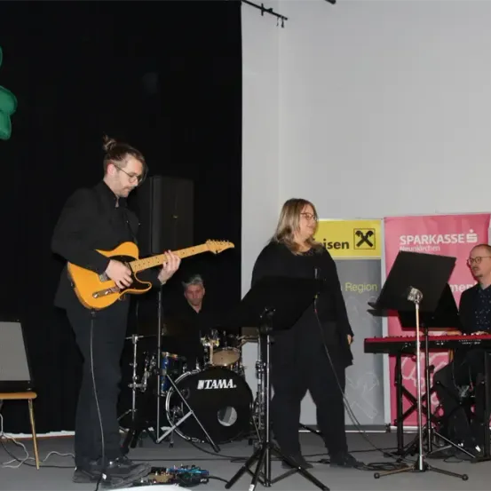 Eine Gruppe Musiker tritt auf der Bühne auf. Ein Mann spielt Gitarre, eine Frau singt und ein anderer Mann spielt Klavier. Ein Schlagzeug und Lautsprecher sind ebenfalls vorhanden.