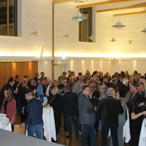 Eine große Indoor-Versammlung mit vielen Menschen in formeller Kleidung, die in einem gut beleuchteten Raum mit hängenden Lampen und Tischen stehen und sich unterhalten.