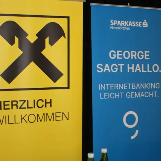 Zwei Banner nebeneinander. Das linke ist gelb mit einem schwarzen gekreuzten Schraubenschlüssel-Symbol und sagt Herzlich Willkommen. Das rechte ist blau und sagt George sagt Hallo. Internetbanking leicht gemacht.