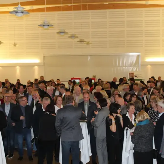 Eine große Gruppe formell gekleideter Personen versammelt sich in einem Konferenzsaal, plaudert und unterhält sich. Tische mit weißen Tischdecken sind in der Mitte aufgestellt. Hängende Lampen beleuchten den Raum.