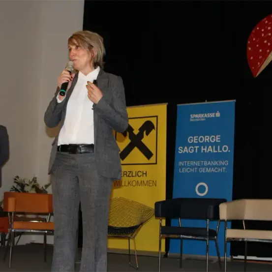 Eine Frau spricht in ein Mikrofon auf der Bühne. Ein Mann steht hinter ihr, und eine Pflanze ist neben ihm. Zwei Banner stehen hinter den Stühlen.