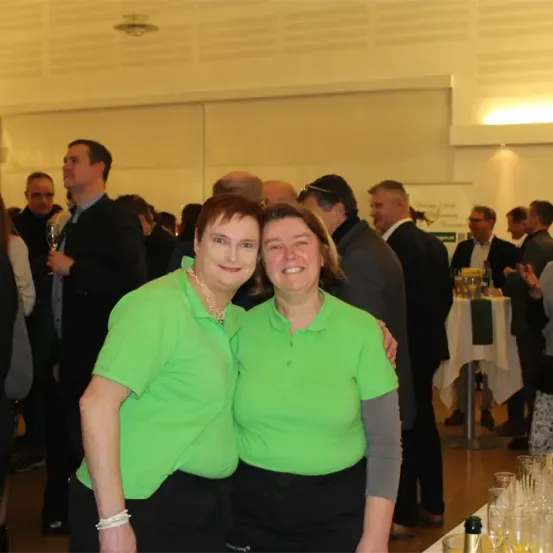 Zwei Frauen in grünen Uniformen stehen eng beieinander auf einer Veranstaltung, lächeln und sind von anderen Menschen umgeben, einige halten Getränke. Dahinter steht ein Tisch, der mit Gläsern und einer Flasche bestückt ist.