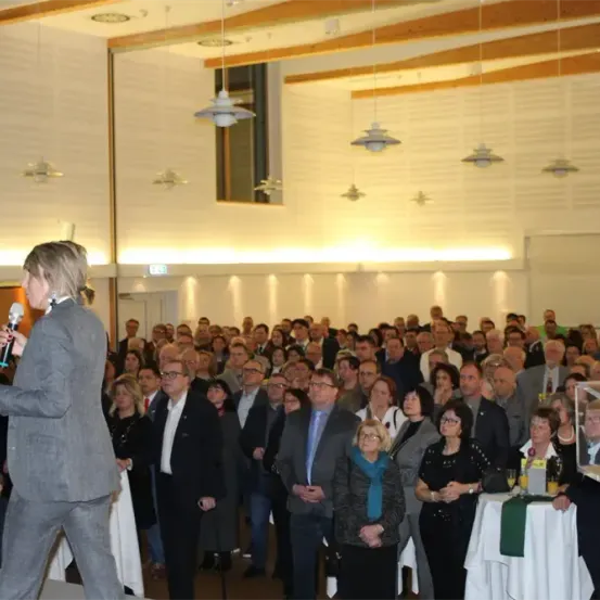 Ein Mann in einem grauen Anzug steht auf der Bühne und spricht zu einem großen Publikum in einem gut beleuchteten Saal. Die formell gekleideten Zuhörer stehen aufmerksam und einige halten Getränke in den Händen.