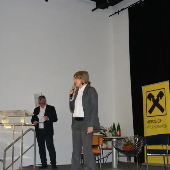 Eine Frau spricht in ein Mikrofon auf der Bühne, während ein Mann in der Nähe ein Blatt Papier liest. Dahinter befindet sich ein gelbes Schild mit einem roten 'X' und den Worten 'Herzlich Willkommen'. Es gibt Stühle und einen Tisch mit Pflanzen.