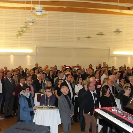 Eine große Gruppe von Menschen ist drinnen versammelt, wahrscheinlich für eine Veranstaltung. Viele sind formell gekleidet, und einige stehen in der Nähe von Tischen. Ein Klavier ist auf der rechten Seite sichtbar.