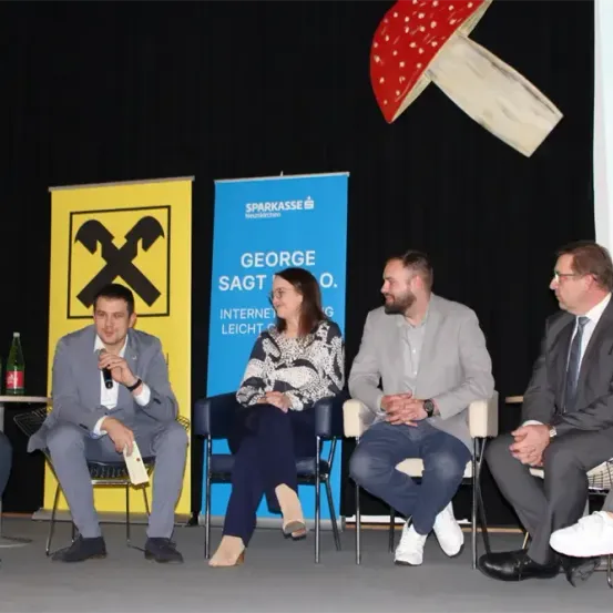 Fünf Personen sitzen in Stühlen vor dem Publikum, eine hält ein Mikrofon. Hinter ihnen stehen zwei Banner, eines mit einem durchgestrichenen X und das andere mit dem Text 'George sagt'. An der Wand befindet sich ein übergroßer roter Pilz.