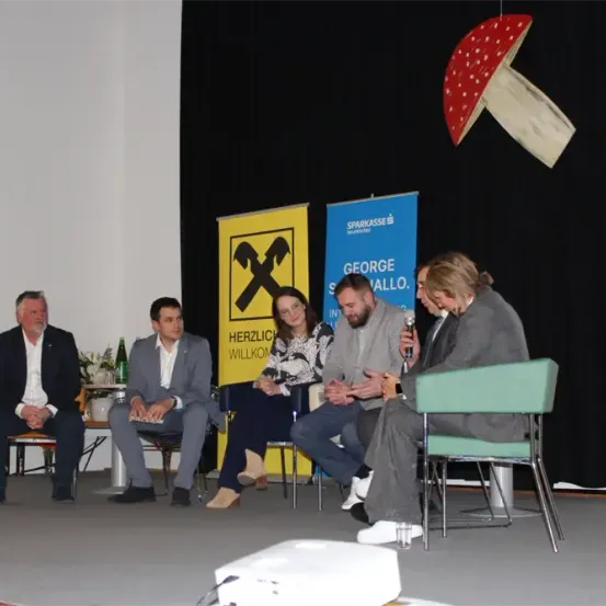 Fünf Personen sitzen auf Stühlen auf einer Bühne und führen eine Diskussion. Zwei Banner hängen an der Wand hinter ihnen. Eines der Schilder lautet 'Herzlich Willkommen' und das andere zeigt ein Logo.