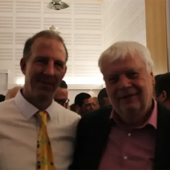 Zwei Männer stehen nah beieinander und lächeln bei einer Zusammenkunft. Der Mann auf der linken Seite trägt eine gemusterte Krawatte, und der Mann auf der rechten Seite trägt ein rosa Hemd. Ein verschwommener Hintergrund zeigt eine Menschenmenge.