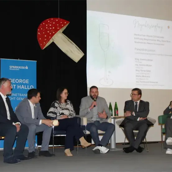 Fünf Personen sitzen auf Stühlen auf einer Bühne mit einem großen Pilz-Hintergrund. Ein Mann hält ein Mikrofon und spricht, während andere zuhören. Eine Projektionsleinwand hinter ihnen zeigt Text und Bilder.