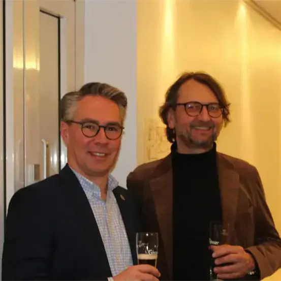 Zwei Männer stehen eng nebeneinander und lächeln. Beide halten Gläser in den Händen, links ein Getränk und rechts ein Bier. Sie befinden sich in einem Flur.