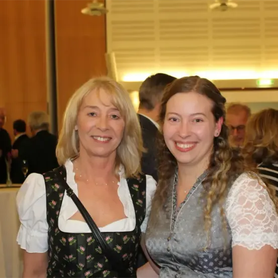 Zwei Frauen, eine in traditioneller bayerischer Kleidung und die andere in einem Spitzenoberteil, lächeln für ein Foto vor einem verblassten Hintergrundpublikum.