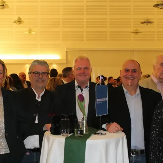 Fünf Menschen lächeln für die Kamera, zwei Frauen und drei Männer, bei einer formellen Veranstaltung, mit einem Tisch im Vordergrund.