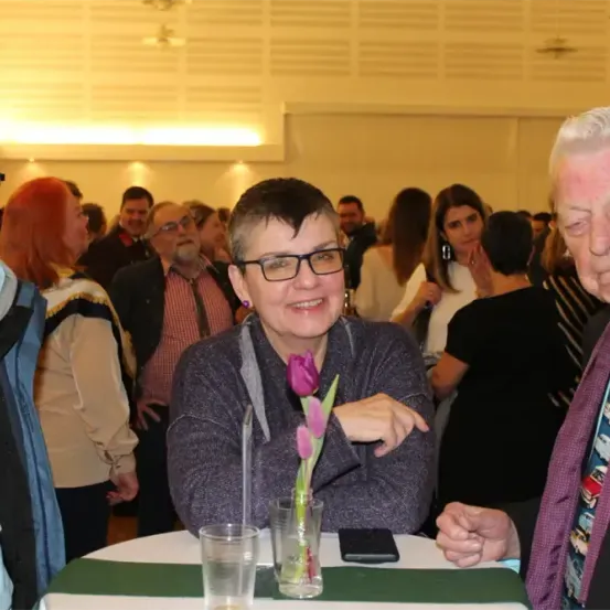 Eine Gruppe von Menschen bei einer Veranstaltung, eine Frau mit Brille lächelt und zeigt auf eine Blume. Ein älterer Mann mit einem gemusterten Krawatte steht in der Nähe.