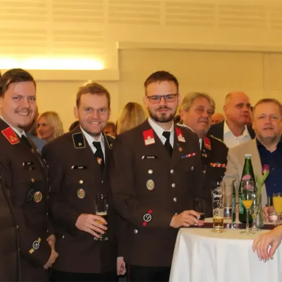 Eine Gruppe von Männern in Militäruniformen steht bei einer Veranstaltung zusammen und hält Getränke in den Händen. Sie scheinen zu lächeln und unterhalten sich. Im Vordergrund ist ein Tisch mit Getränken und einer Blumenarrangement zu sehen.