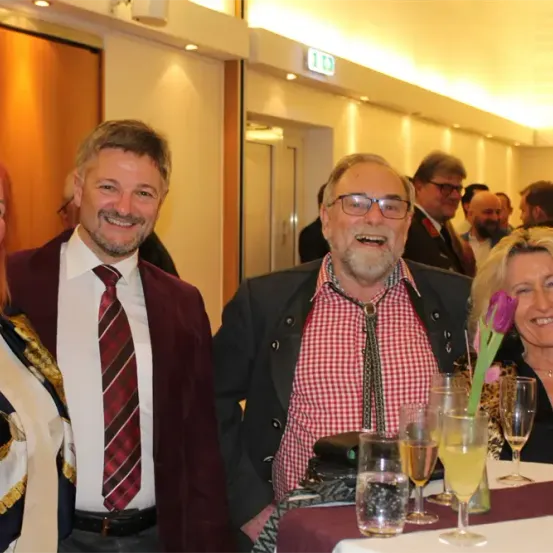 Vier Erwachsene sind an einem Tisch versammelt, lächeln und posieren für ein Foto. Auf dem Tisch befinden sich Gläser und eine Blume, und im Hintergrund stehen weitere Personen.