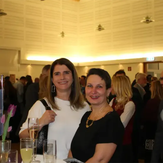 Zwei Frauen lächeln für die Kamera bei einer formellen Veranstaltung. Eine hält ein Glas Champagner. Im Hintergrund unterhalten sich mehrere Personen.