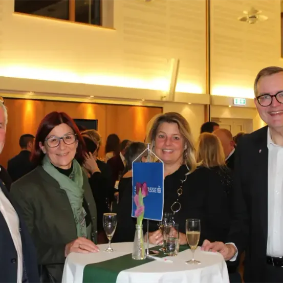 Vier Menschen lächeln für ein Foto bei einer gesellschaftlichen Veranstaltung, mit Weinläsern und einem Blumengesteck auf einem Tisch vor ihnen.