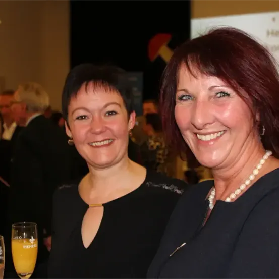 Zwei Frauen in schwarzen Kleidern lächeln bei einer gesellschaftlichen Veranstaltung, eine mit einem Glas Orangensaft und die andere mit einer Perlenkette. Dahinter sind ein Mann im Anzug und andere verschwommen.