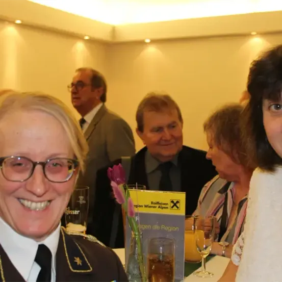 Eine Gruppe von Menschen sitzt lächelnd am Tisch. Eine Frau trägt eine Brille und eine Uniform. Hinter ihnen unterhalten sich zwei Männer in Anzügen und Krawatten. Auf dem Tisch liegt ein gelber Prospekt und eine Blume in einer Vase.