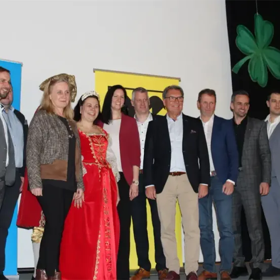 Eine Gruppe von Menschen steht gemeinsam vor einer weißen Wand mit einem gelben Banner und einem grünen Kleeblatt. Die Personen sind in verschiedenen Kleidungsstücken gekleidet, darunter Anzüge, Kleider und eine Krone.