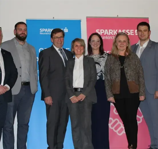 Eine Gruppe von Menschen, die formell gekleidet sind, steht gemeinsam vor zwei Bannern. Ein Banner ist blau und das andere ist rosa, mit Text darauf.