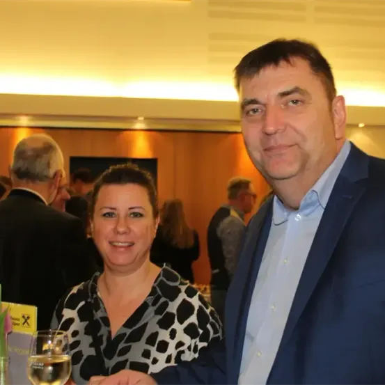 Ein Mann und eine Frau lächeln zur Kamera bei einer formellen Veranstaltung. Hinter ihnen stehen und plaudern andere Leute. Der Mann trägt einen marineblauen Anzug und die Frau ein schwarz-weißes Oberteil.