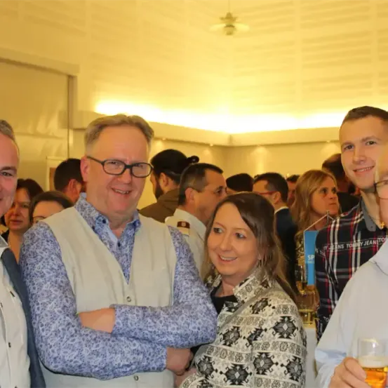 Mehrere Personen lächeln und posieren für ein Foto in einer Innenumgebung. Ein Mann hält ein Glas Bier. Eine Person steht neben einer Glasflasche.