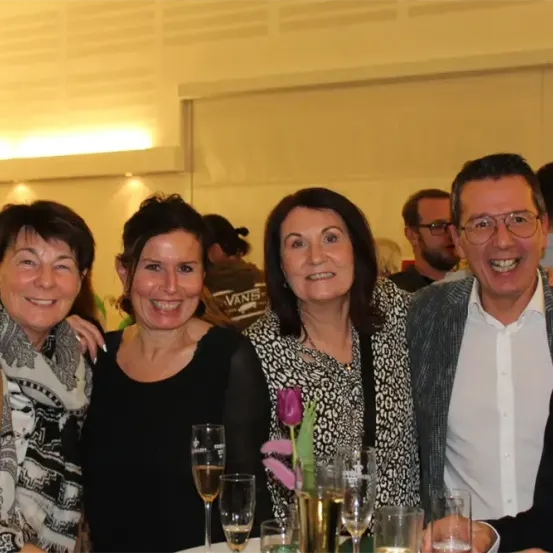 Eine Gruppe von Menschen auf einer Party, lächelnd, mit einem Mann in einem weißen Hemd, zwei Frauen in Schwarz und einer Frau in einem Leopardendruckkleid, die Weingläser halten, und einem Mann mit Brille.