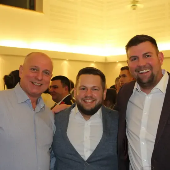 Drei Männer stehen bei einem gesellschaftlichen Ereignis zusammen und lächeln für die Kamera. Der Mann links ist kahlköpfig, der in der Mitte hat einen Bart und der Mann rechts trägt einen Anzug.