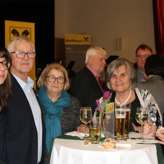 Eine Gruppe von Menschen, die lächelnd und für ein Foto posieren, um einen Tisch mit Weingläsern und Essen. Ein Mann mit Brille und eine Frau mit einem Schal sind in der Mitte.