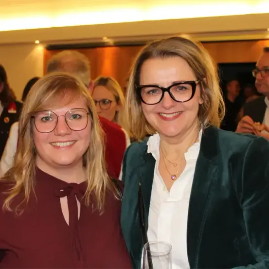 Zwei Frauen lächeln und posieren für ein Foto bei einer Veranstaltung. Die Frau auf der linken Seite trägt eine Brille und eine kastanienbraune Bluse. Die Frau auf der rechten Seite trägt einen grünen Blazer und eine Halskette. Hinter ihnen unterhalten sich Menschen und einige essen. Der Raum ist von Deckenleuchten beleuchtet.