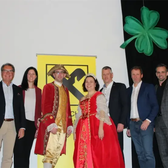 Eine Gruppe von Menschen steht vor einem gelben Banner mit einem Mann und einer Frau in historischen Kostümen. Sie scheinen für ein Foto zu posieren.