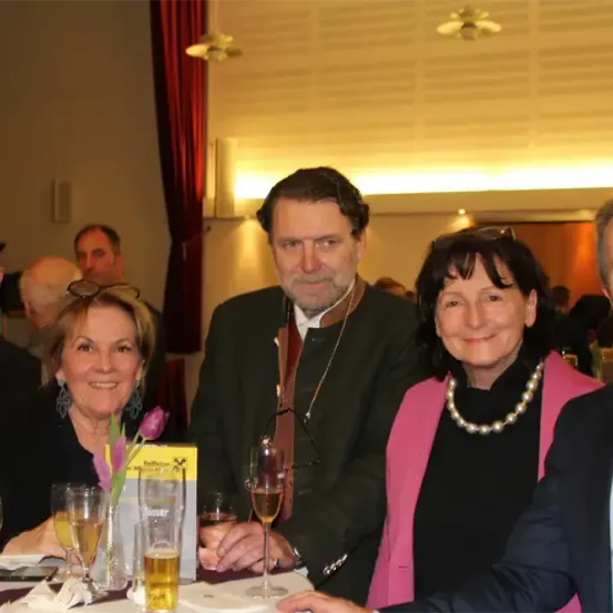 Fünf Personen, drei Männer und zwei Frauen, sitzen lächelnd an einem Tisch in einem Bankettsaal. Auf dem Tisch stehen Getränke und Gläser, wobei eine Frau eine Blume hält. Im Hintergrund befinden sich Deckenventilatoren und ein roter Vorhang.