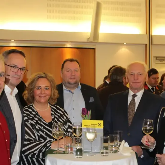 Gruppe formell gekleideter Männer und Frauen, die bei einem gesellschaftlichen Ereignis zusammenstehen und für die Kamera lächeln. Ein Tisch mit Weinläsern und einem gelben Banner im Vordergrund.