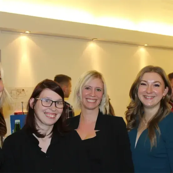 Fünf Frauen stehen zusammen und lächeln in die Kamera. Sie tragen formelle Kleidung. Eine hat eine Brille und eine andere hat lange Haare. Im Hintergrund sind zwei Männer zu sehen.