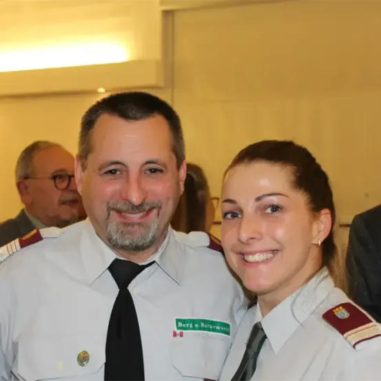 Ein Mann und eine Frau in Uniformen, beide lächelnd, stehen bei einer Veranstaltung eng beieinander. Der Mann hat ein Abzeichen und ein grünes Abzeichen, während die Frau ein rotes Abzeichen auf der Schulter hat. Verschwommene Menschen stehen hinter ihnen.