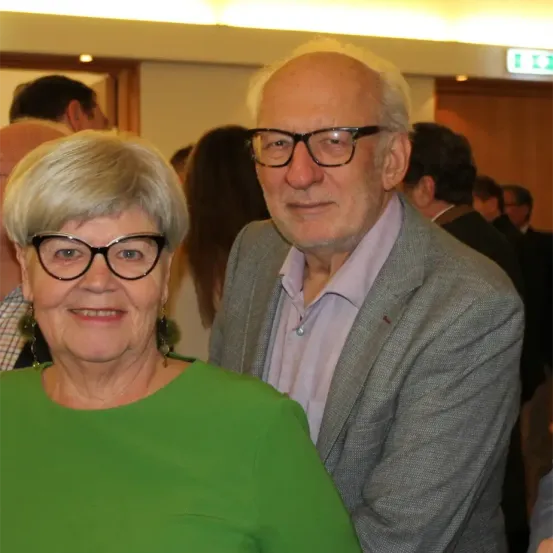 Ein älteres Paar, beide mit Brille, lächelt zur Kamera bei einer formellen Veranstaltung. Dahinter stehen und unterhalten sich Menschen.