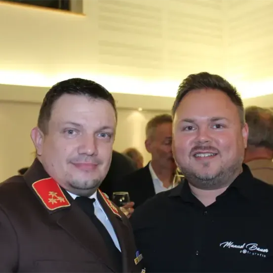 Zwei Männer stehen eng beieinander, lächeln, mit einem verschwommenen Hintergrund von Menschen und einer weißen Decke.