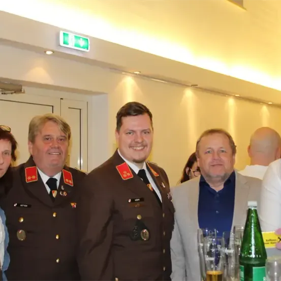 Eine Gruppe von Menschen in Uniformen und Anzügen posiert für ein Foto in einem Gebäude. Auf einem Tisch stehen Gläser, eine Flasche und andere Gegenstände. Eine Ausfahrtstafel ist an der Decke angebracht.